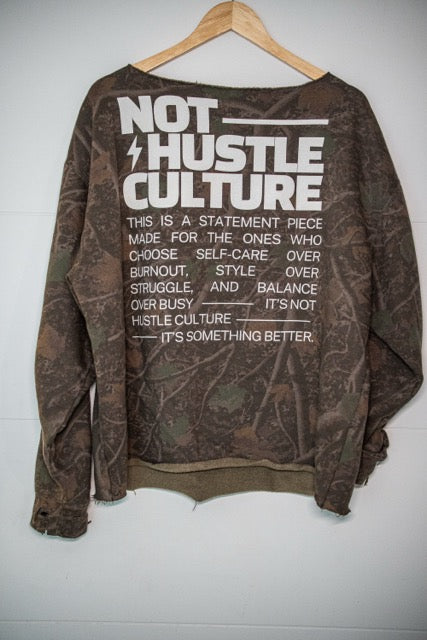 A brown camouflage long-sleeve sweatshirt with a large white graphic text print on the back, hanging on a wooden hanger against a white wall.