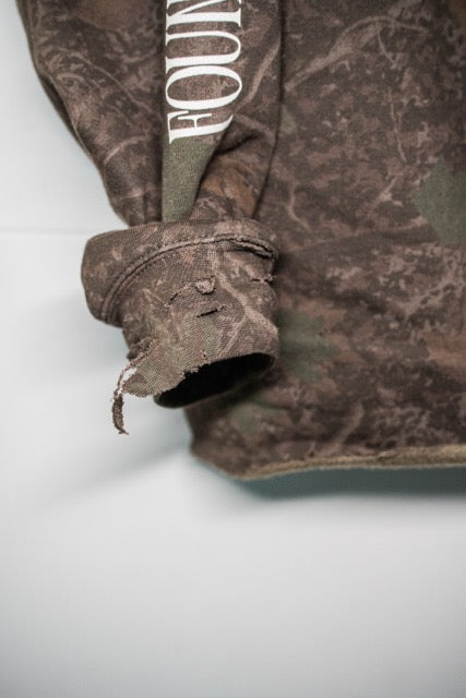 Close-up of a camouflage-patterned garment, likely a hoodie or sweatshirt, showing a rolled-up sleeve with a frayed hem and white text reading 'FOUN' on the shoulder area.