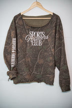 A long-sleeve camouflage top hanging on a wooden hanger against a white paneled wall. The shirt features a brown and green woodland camo pattern with white text reading 'SPORTS HUSTlers CLUB' on the front and 'FOUNDRING MEMBER' printed down one sleeve.