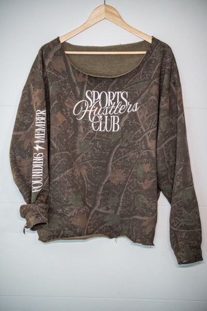 A long-sleeve camouflage top hanging on a wooden hanger against a white paneled wall. The shirt features a brown and green woodland camo pattern with white text reading 'SPORTS HUSTlers CLUB' on the front and 'FOUNDRING MEMBER' printed down one sleeve.