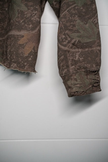 Close-up of a camouflage-patterned garment, likely a jacket or sweatshirt, showing the sleeve cuff with frayed edges and a distressed finish, hanging against a light gray wall.