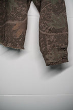 Close-up of a camouflage-patterned garment, likely a jacket or sweatshirt, showing the sleeve cuff with frayed edges and a distressed finish, hanging against a light gray wall.