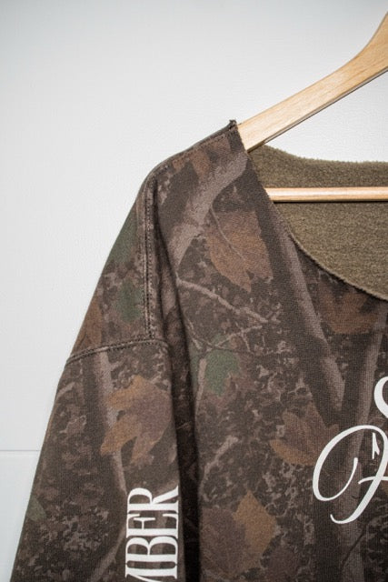 A close-up of a camouflage-patterned long-sleeve shirt hanging on a wooden hanger against a plain light gray wall. The shirt features a brown and green camo print with white script lettering on the chest and sleeve.
