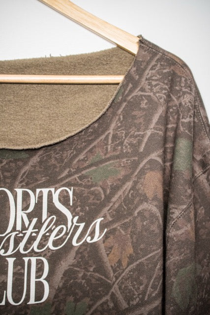 A close-up of a camouflage-patterned long-sleeve shirt hanging on a wooden hanger, featuring a scoop neck with a tan fleece lining and white text reading 'SPORTS Wrestlers CLUB'.