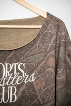 A close-up of a camouflage-patterned long-sleeve shirt hanging on a wooden hanger, featuring a scoop neck with a tan fleece lining and white text reading 'SPORTS Wrestlers CLUB'.