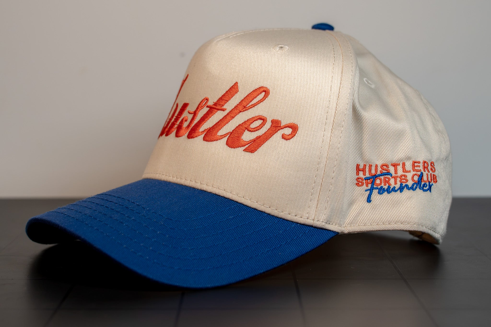 angled view of the cream and royal blue hustler script cap showing founder embroidery on the side panel